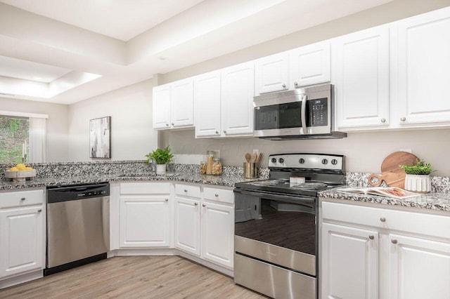 kitchen with light stone countertops, appliances with stainless steel finishes, light wood-type flooring, a tray ceiling, and white cabinets