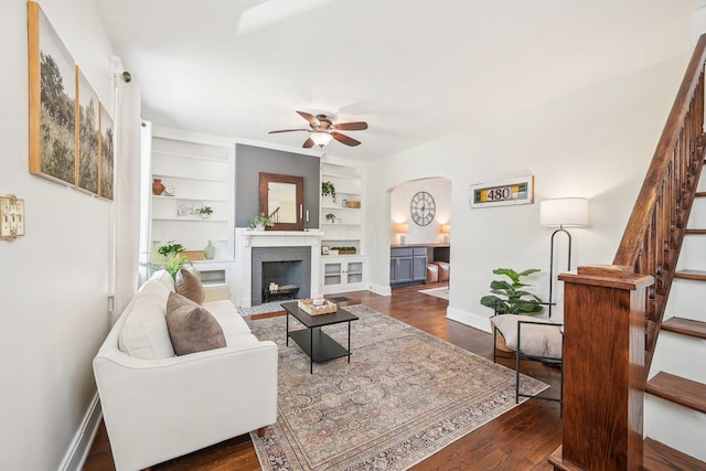 living room with built in features, baseboards, dark wood-style floors, arched walkways, and a ceiling fan
