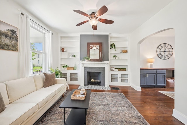 living room with a ceiling fan, built in features, arched walkways, a fireplace, and dark wood-type flooring
