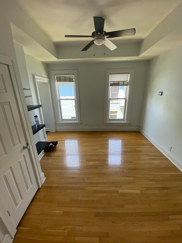 empty room featuring ceiling fan, a healthy amount of sunlight, a tray ceiling, and light hardwood / wood-style flooring