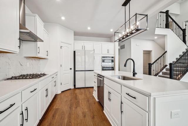 kitchen with sink, wall chimney range hood, an island with sink, white cabinets, and appliances with stainless steel finishes