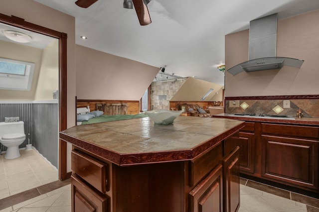 kitchen with tile countertops, stainless steel gas stovetop, wall chimney range hood, light tile patterned floors, and a kitchen island