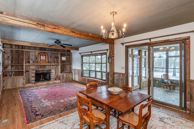 dining room with ceiling fan with notable chandelier, built in features, wooden walls, light hardwood / wood-style floors, and a stone fireplace