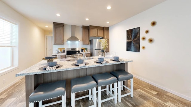 kitchen featuring appliances with stainless steel finishes, a large island, wall chimney range hood, hardwood / wood-style flooring, and a breakfast bar area