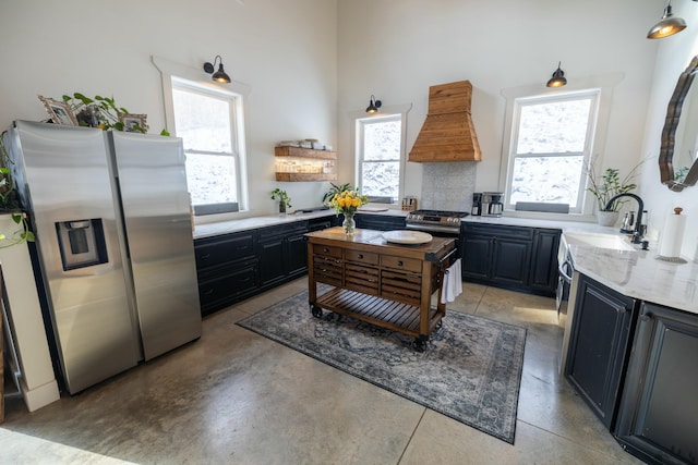 kitchen with light stone countertops, sink, premium range hood, stainless steel fridge, and a towering ceiling