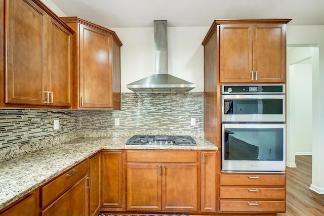 kitchen featuring backsplash, wall chimney exhaust hood, light hardwood / wood-style floors, light stone counters, and stainless steel appliances