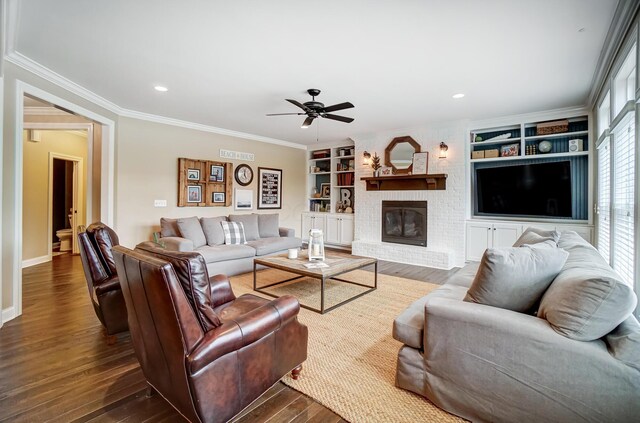 living room with built in features, dark wood finished floors, ceiling fan, crown molding, and a brick fireplace