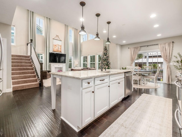 kitchen featuring stainless steel dishwasher, light stone counters, decorative light fixtures, a center island with sink, and white cabinetry