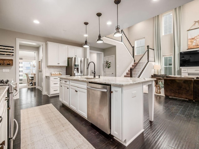 kitchen with hanging light fixtures, stainless steel appliances, light stone counters, a center island with sink, and white cabinets