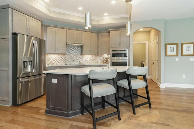 kitchen with decorative backsplash, stainless steel appliances, a raised ceiling, decorative light fixtures, and a center island with sink