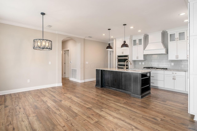 kitchen featuring pendant lighting, white cabinets, premium range hood, and an island with sink