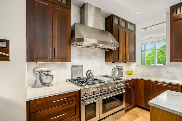 kitchen with backsplash, double oven range, wall chimney exhaust hood, and light stone counters