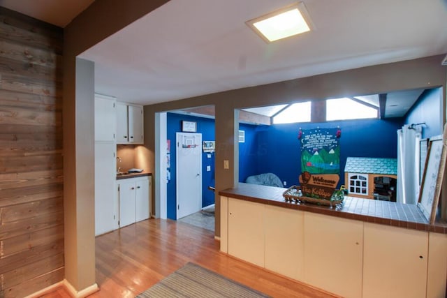 kitchen with tile countertops, white cabinets, wooden walls, light wood-type flooring, and kitchen peninsula