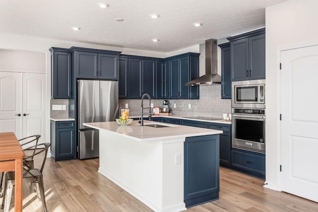 kitchen with stainless steel appliances, sink, light hardwood / wood-style floors, an island with sink, and wall chimney range hood