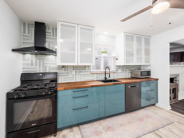 kitchen with sink, wall chimney range hood, a stone fireplace, butcher block countertops, and appliances with stainless steel finishes