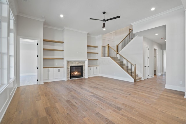 unfurnished living room featuring a stone fireplace, ceiling fan, crown molding, light wood-type flooring, and built in shelves