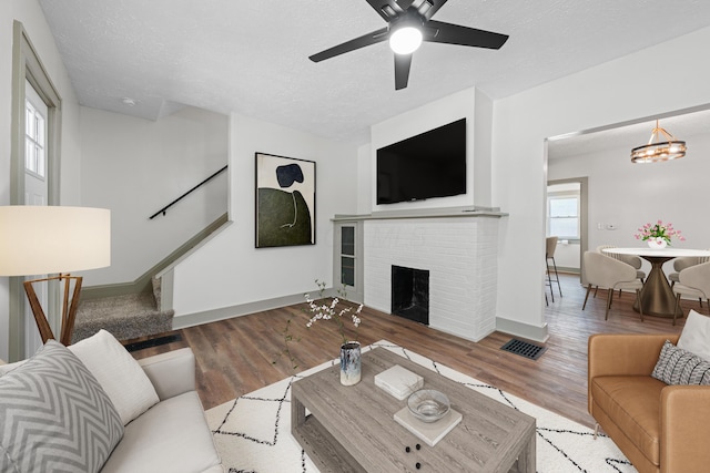 living room with ceiling fan with notable chandelier, light wood-type flooring, a textured ceiling, and a brick fireplace