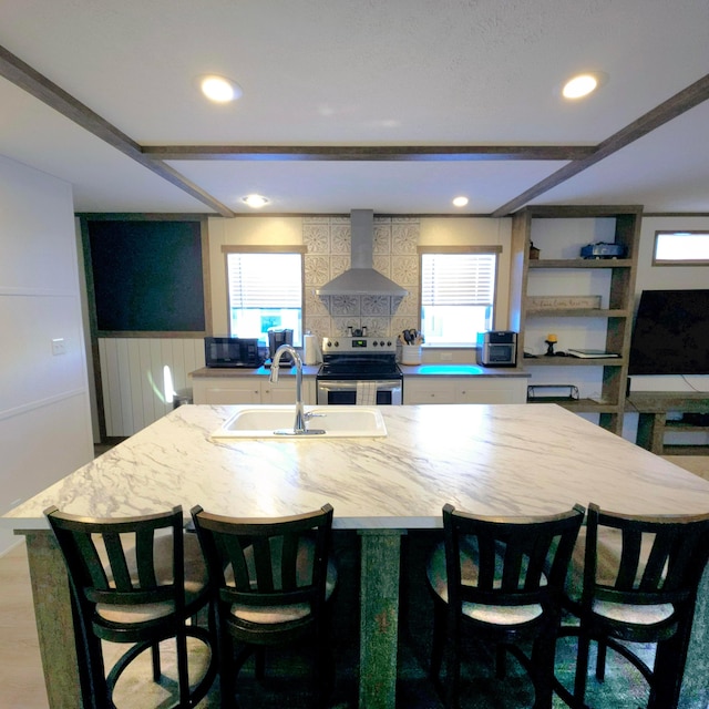 kitchen featuring stainless steel electric range oven, a kitchen island with sink, sink, and wall chimney exhaust hood