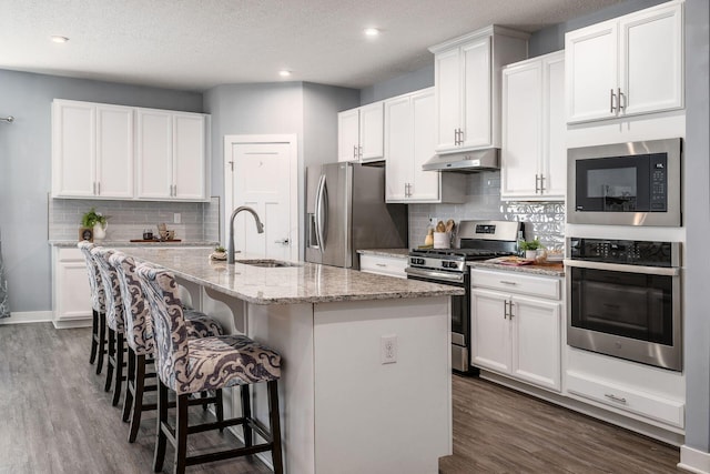 kitchen with sink, white cabinetry, stainless steel appliances, light stone countertops, and an island with sink