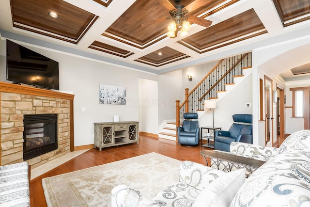 living room featuring coffered ceiling, a stone fireplace, wood-type flooring, and ornamental molding