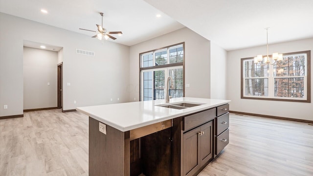 kitchen featuring an island with sink, light hardwood / wood-style floors, ceiling fan with notable chandelier, dark brown cabinets, and sink