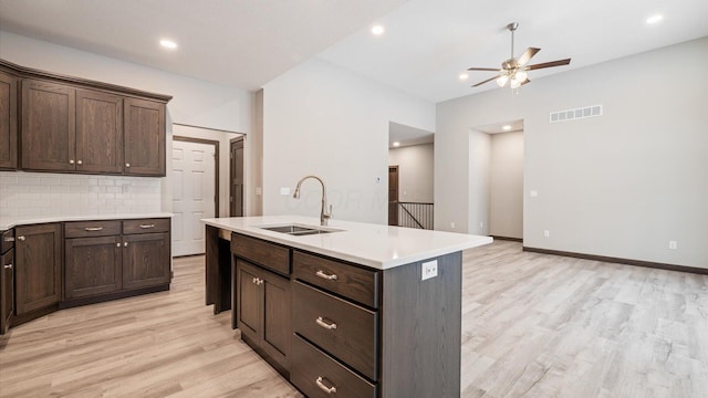 kitchen featuring sink, light wood-type flooring, backsplash, dark brown cabinets, and a kitchen island with sink