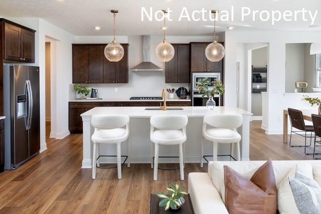 kitchen featuring decorative light fixtures, an island with sink, dark brown cabinets, wall chimney range hood, and appliances with stainless steel finishes