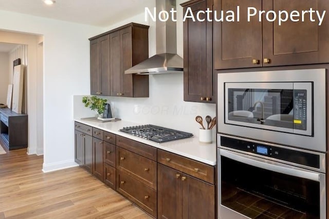 kitchen featuring stainless steel appliances, light wood-type flooring, wall chimney range hood, and dark brown cabinetry