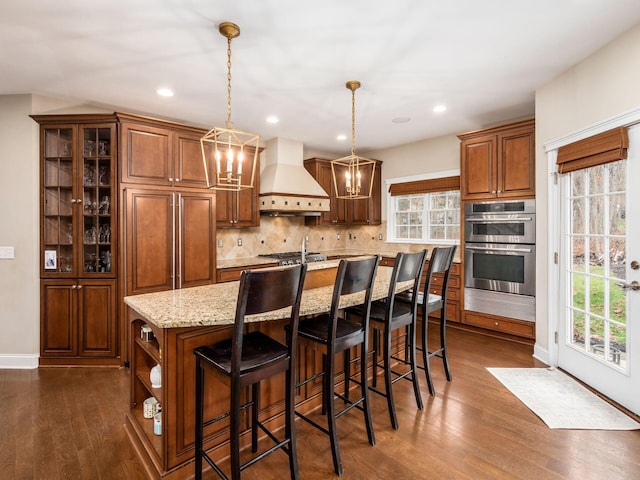 kitchen with custom exhaust hood, hanging light fixtures, an island with sink, stainless steel appliances, and decorative backsplash