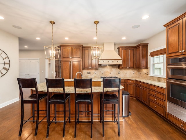 kitchen featuring stainless steel double oven, an inviting chandelier, a kitchen island with sink, and custom exhaust hood