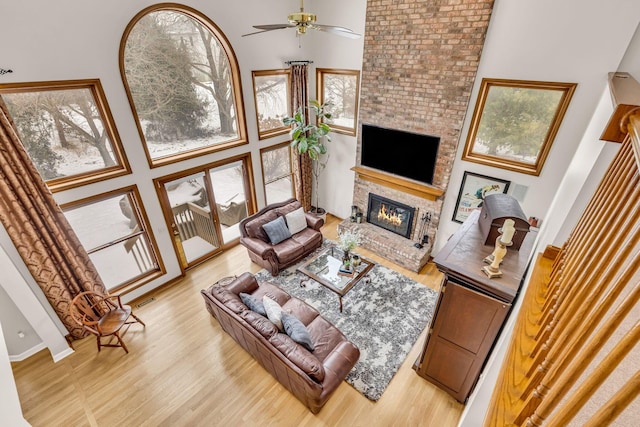 living room with ceiling fan, light hardwood / wood-style flooring, a towering ceiling, and a brick fireplace