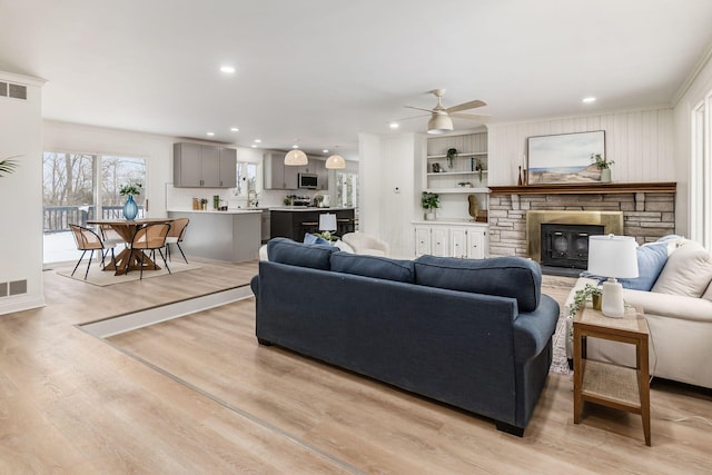 living room featuring a stone fireplace, ceiling fan, light wood-type flooring, crown molding, and built in shelves