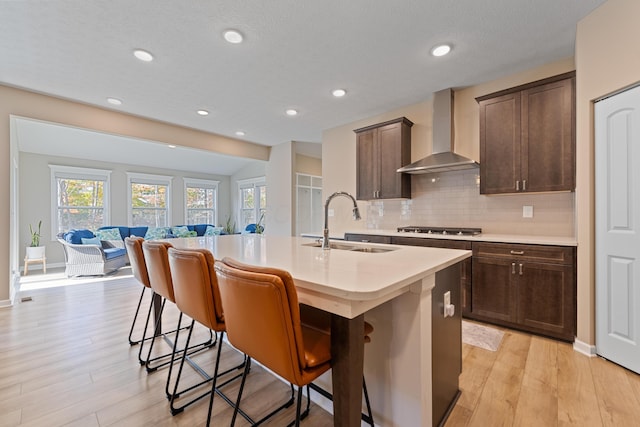 kitchen with wall chimney range hood, an island with sink, sink, a kitchen breakfast bar, and light hardwood / wood-style flooring