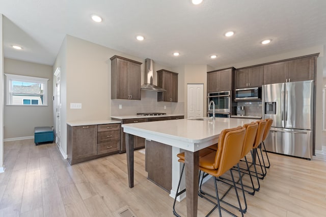 kitchen featuring appliances with stainless steel finishes, wall chimney range hood, light wood-type flooring, a center island with sink, and a breakfast bar area