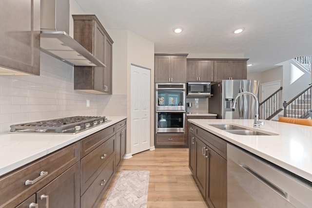 kitchen with decorative backsplash, sink, light wood-type flooring, appliances with stainless steel finishes, and wall chimney exhaust hood