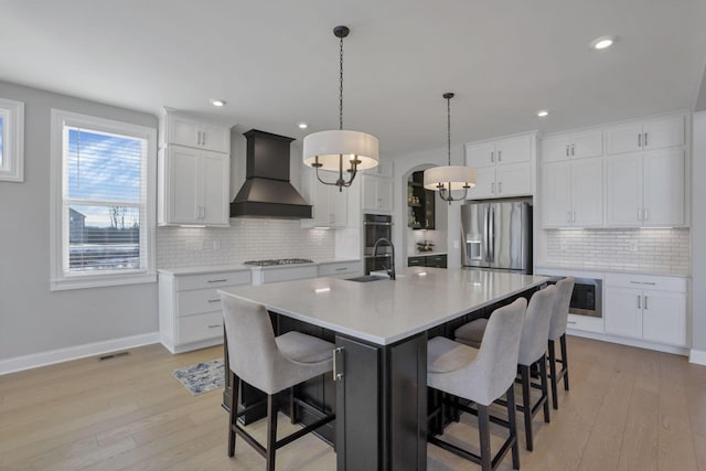 kitchen with pendant lighting, white cabinets, stainless steel appliances, custom range hood, and a center island with sink