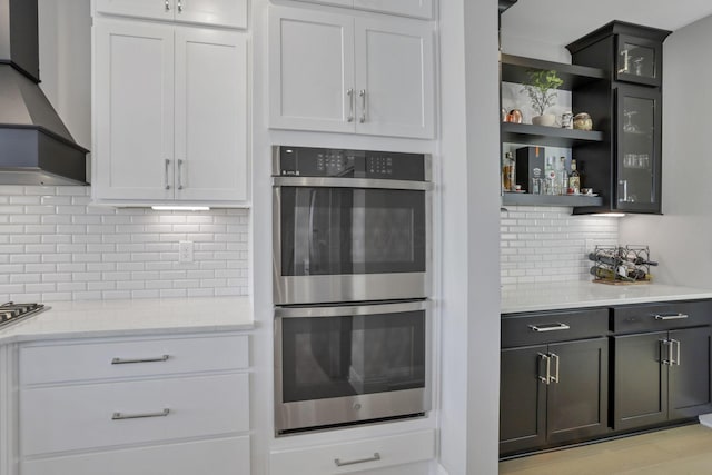 kitchen with custom exhaust hood, white cabinetry, light stone countertops, and double oven