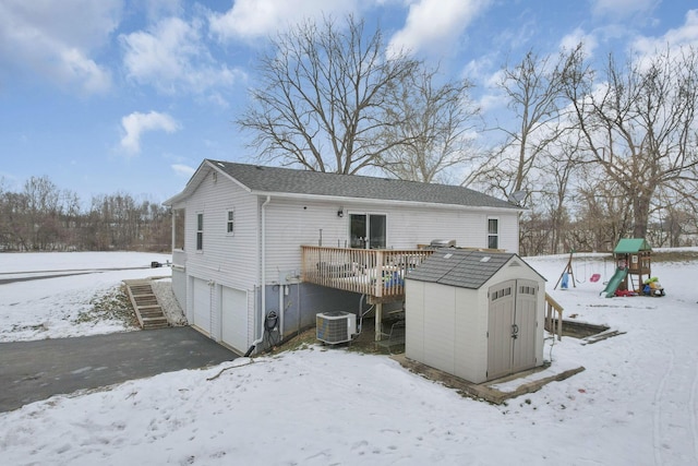 snow covered property featuring a playground, a wooden deck, a garage, central AC unit, and a shed