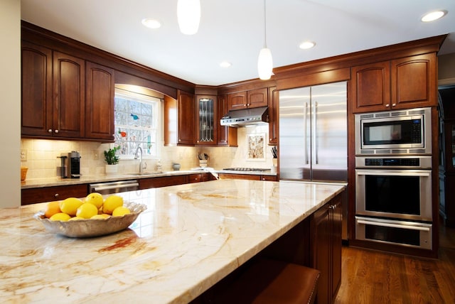 kitchen with built in appliances, light stone counters, under cabinet range hood, dark wood-type flooring, and decorative backsplash