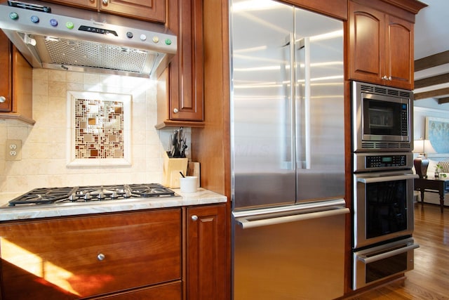 kitchen with backsplash, brown cabinets, under cabinet range hood, and built in appliances