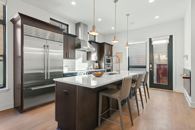 kitchen featuring decorative light fixtures, an island with sink, backsplash, stainless steel appliances, and wall chimney exhaust hood