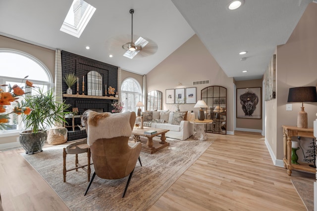 living room featuring a skylight, high vaulted ceiling, light wood-type flooring, and a brick fireplace