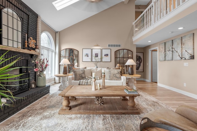 living room with a brick fireplace, high vaulted ceiling, light hardwood / wood-style floors, and a skylight