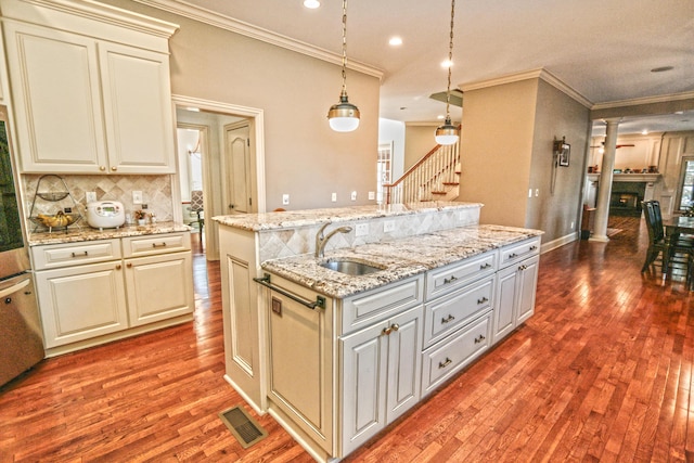 kitchen featuring ornate columns, decorative light fixtures, a kitchen island with sink, light stone counters, and sink