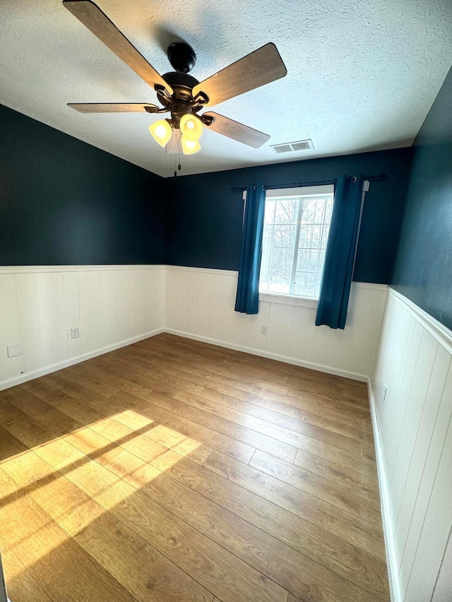 unfurnished room with a textured ceiling, a wainscoted wall, visible vents, a ceiling fan, and wood-type flooring