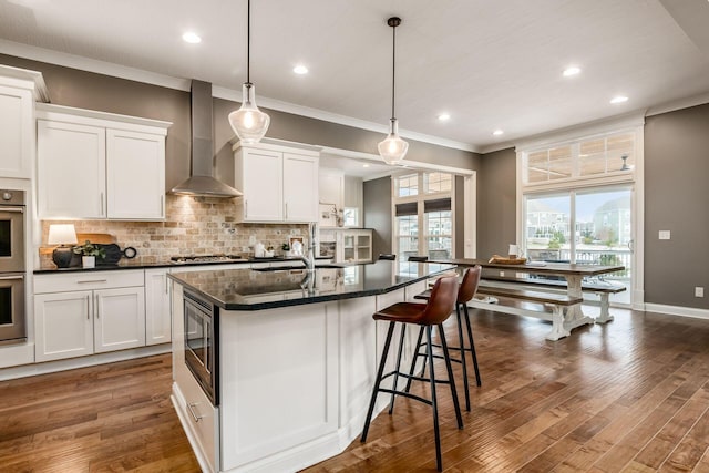 kitchen with appliances with stainless steel finishes, wall chimney exhaust hood, a kitchen island with sink, and white cabinets