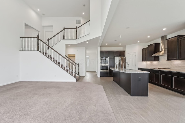 kitchen with sink, tasteful backsplash, stainless steel appliances, a kitchen island with sink, and wall chimney range hood