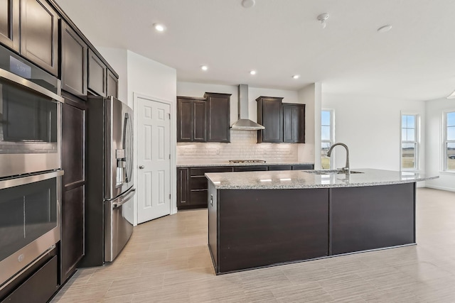 kitchen featuring light stone counters, appliances with stainless steel finishes, wall chimney exhaust hood, and a kitchen island with sink