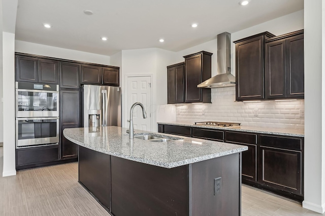 kitchen featuring appliances with stainless steel finishes, sink, wall chimney range hood, light stone countertops, and a center island with sink