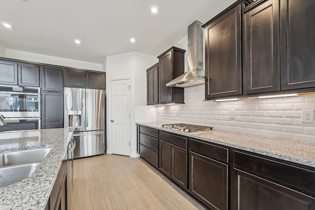 kitchen featuring appliances with stainless steel finishes, tasteful backsplash, sink, light stone countertops, and wall chimney range hood